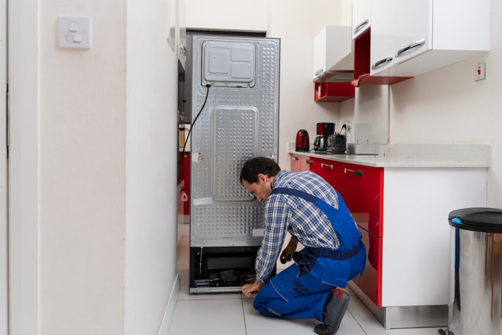 Young Male Serviceman Repairing Broken Refrigerator In Kitchen